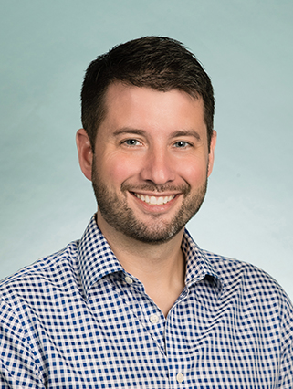 A man with short dark hair, a beard, and wearing a checkered shirt smiles directly at the camera against a plain background.