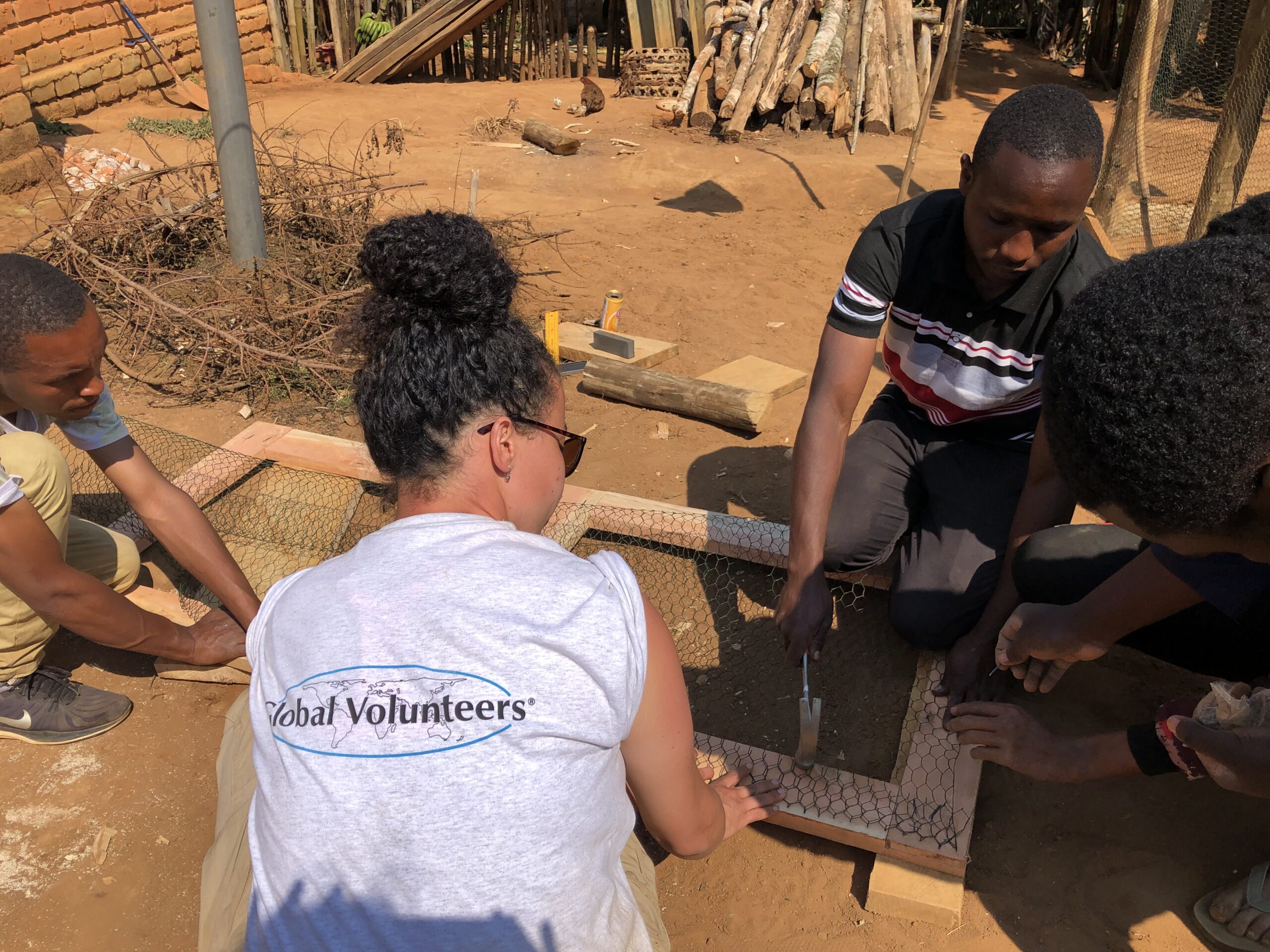 A group of people works together assembling a wire structure outdoors. One person wears a shirt reading "Global Volunteers." Wood, tools, and debris are visible in the background.