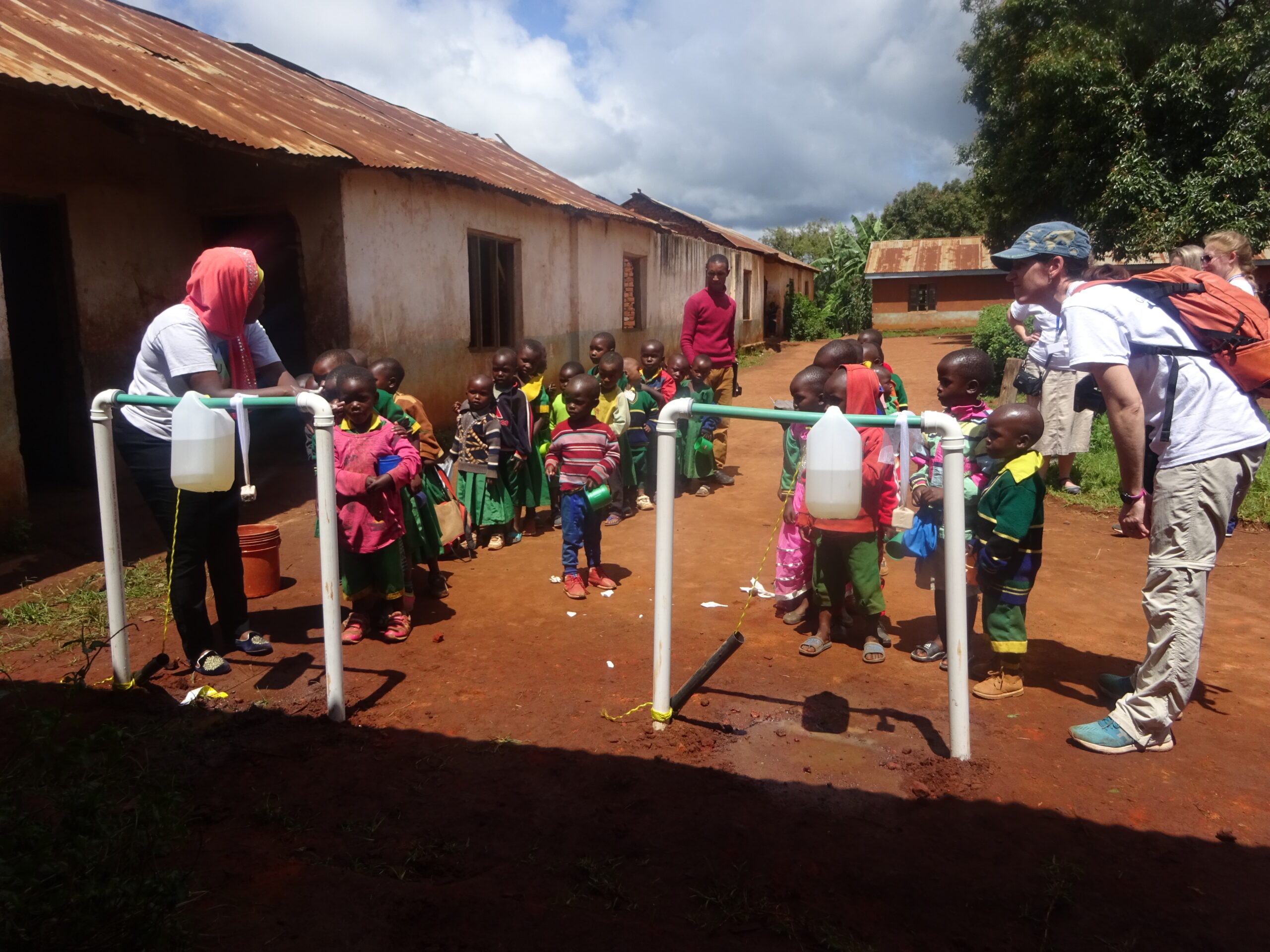 A group of children and two adults gather around makeshift handwashing stations with plastic jugs in an outdoor area near rustic buildings.