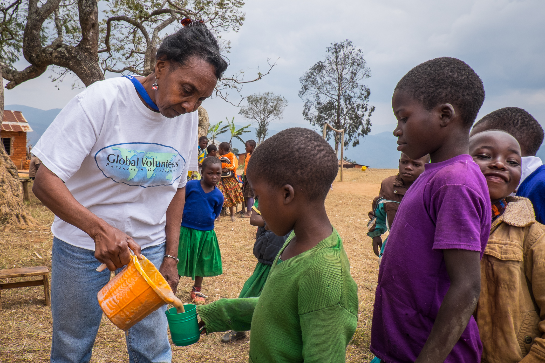 A woman wearing a "Global Volunteers" shirt pours a drink for a child from an orange jug. Several children stand nearby in an outdoor, rural setting.