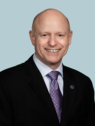 A man in a black suit, light blue dress shirt, and purple tie smiling at the camera against a light blue background.