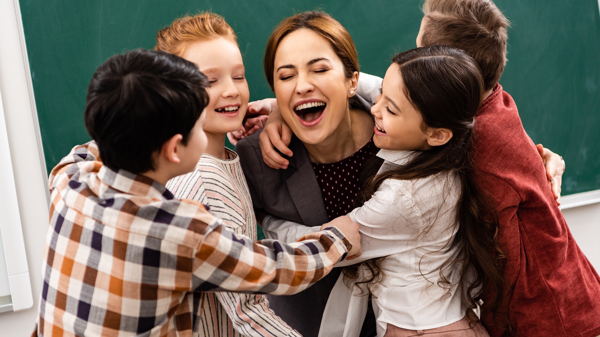 a group of children hugging a teacher in front of a blackboard.