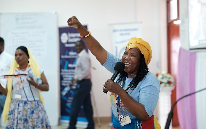 A woman wearing a yellow headscarf and holding a microphone raises her fist in the air while speaking at an indoor event. Other attendees and a whiteboard are visible in the background.