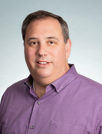 A man with short dark hair, wearing a purple button-down shirt, is looking at the camera with a neutral expression against a light background.