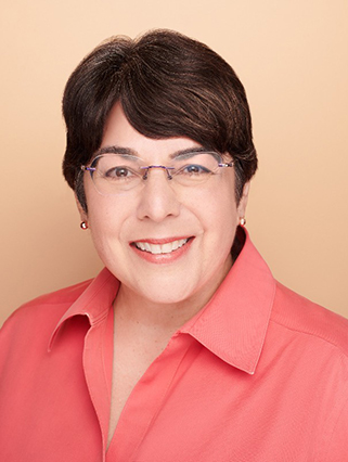 A woman with short dark hair, wearing glasses and a coral-colored shirt, smiles at the camera against a light peach background.