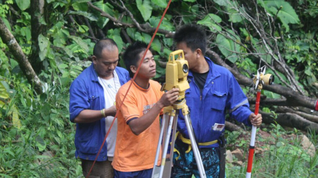 Three men operate surveying equipment in a lush, green forest. One holds a yellow theodolite on a tripod, another supports a red and white staff, and the third adjusts the equipment.