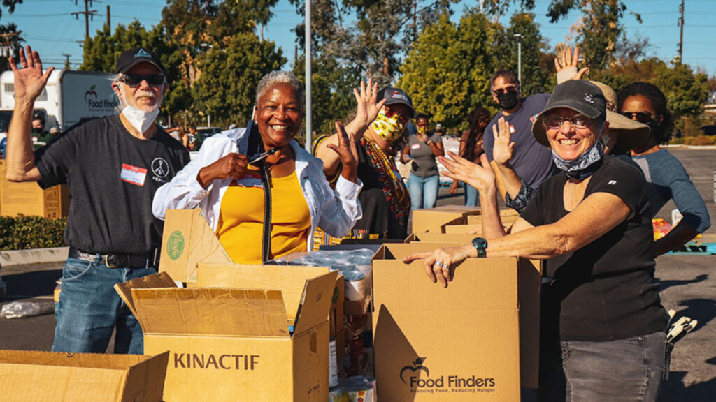 A group of five people standing outdoors, smiling and waving near cardboard boxes labeled "Food Finders." They appear to be participating in a community food distribution event.