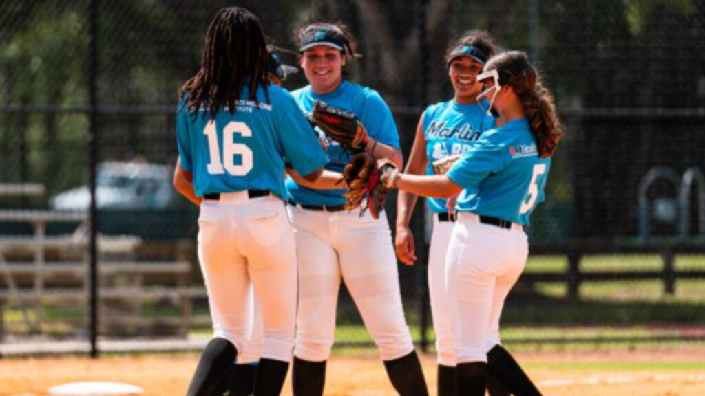 Four softball players in teal jerseys and white pants huddle and smile on a baseball field, with greenery and a fence visible in the background.