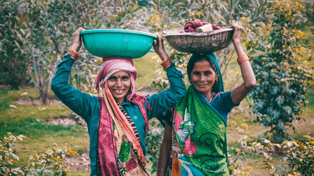 Two women wearing colorful saris and headscarves smile while carrying large bowls on their heads in a lush, green outdoor setting.