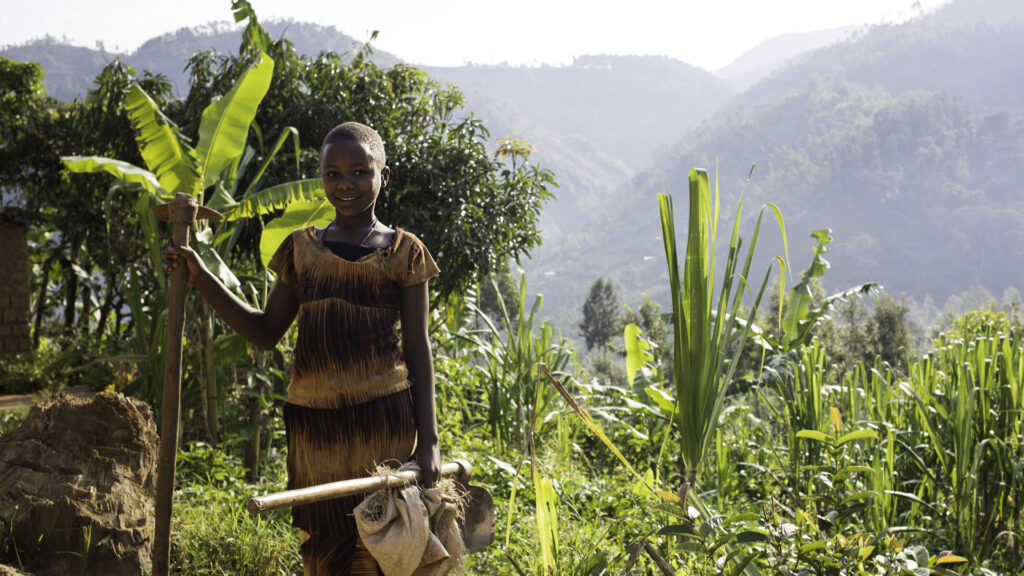 A young person holding farming tools stands in a lush field with hills and trees in the background.