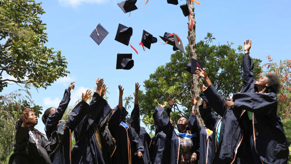 A group of graduates in caps and gowns celebrate by throwing their caps into the air outdoors on a sunny day.