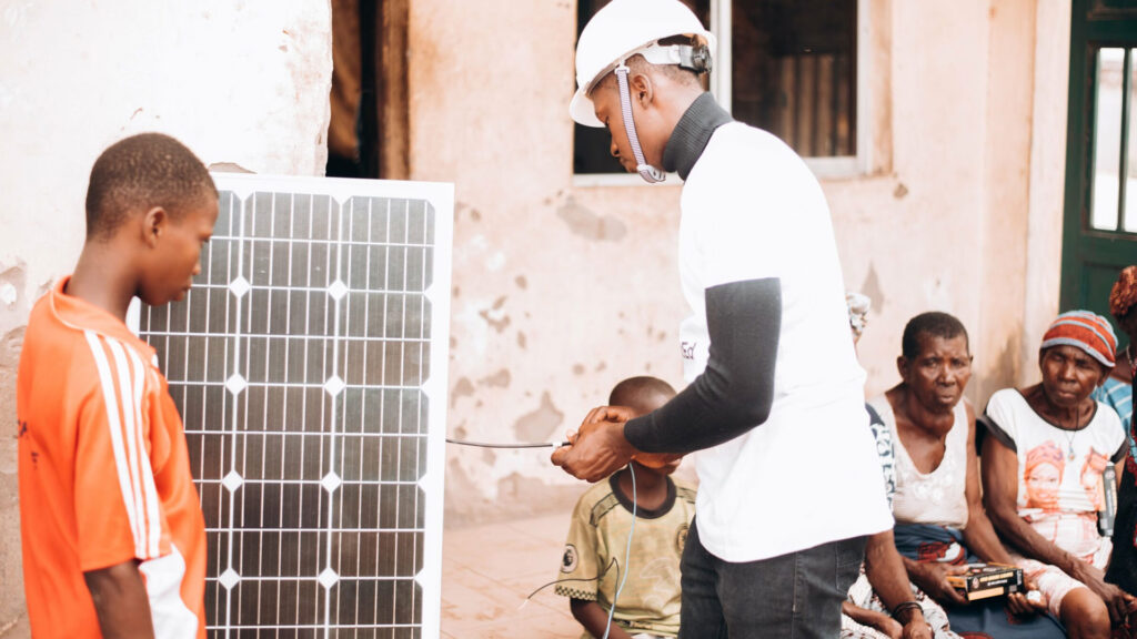 A person in a white hard hat demonstrates a solar panel to a group of people, including children, in an outdoor setting.