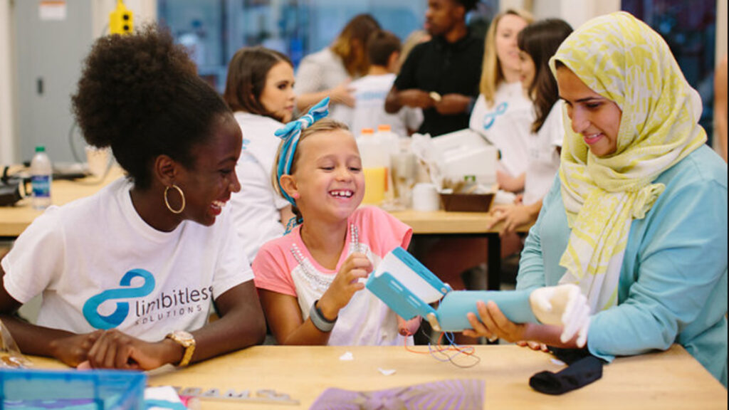 A group of diverse individuals, including a young girl, engage in a workshop setting. Two women assist the girl, one showing her a prosthetic limb. Participants wear "Limbitless Solutions" shirts.