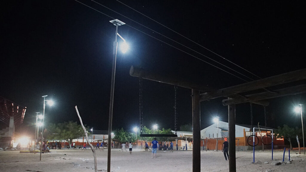 A nighttime scene of an outdoor playground with swings and exercise equipment. The area is illuminated by streetlights, and several people are visible in the background.
