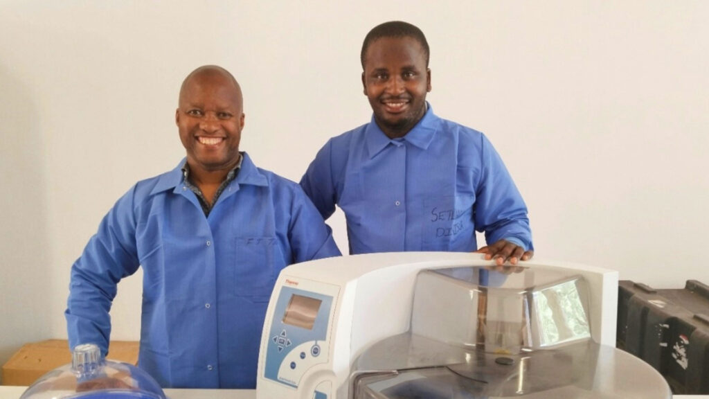 Two men wearing blue lab coats stand smiling beside a large laboratory machine. One has his hand on the machine, and various lab equipment is visible.