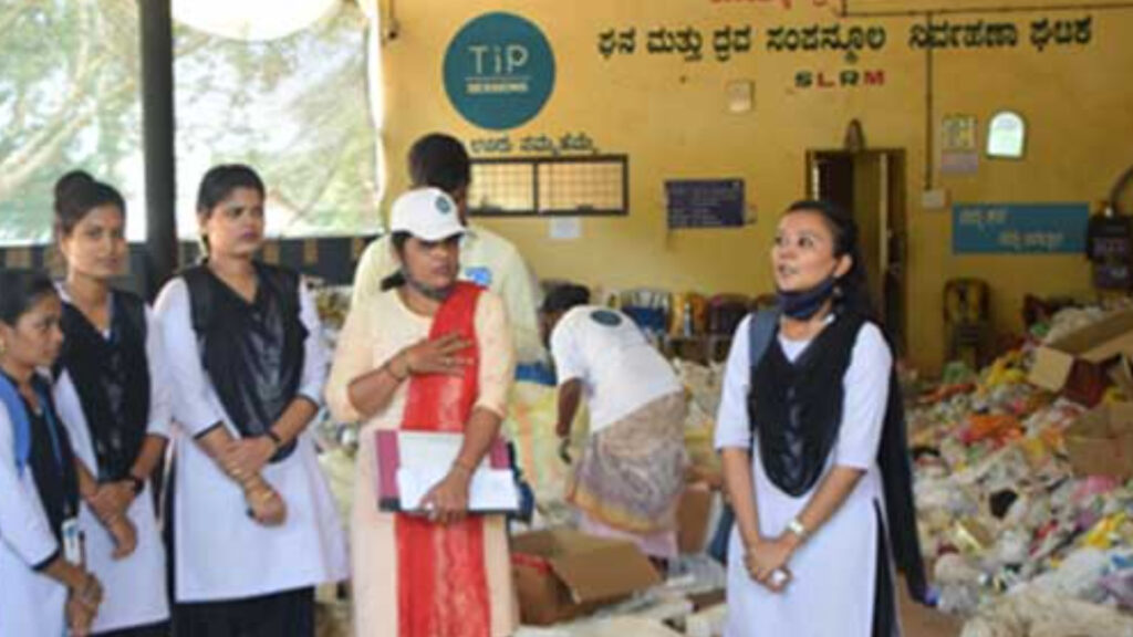 A group of women stands in front of a room filled with sorted waste materials. One woman is addressing the group while others listen. Wall signs in Kannada and English are visible in the background.