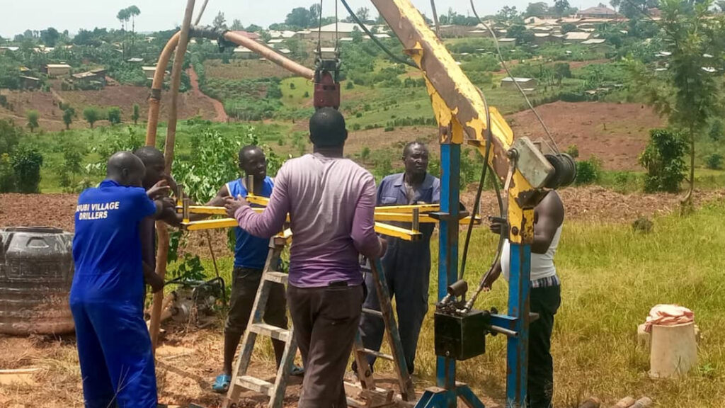People working together on a well-drilling project in a rural area, with green vegetation and scattered houses in the background.
