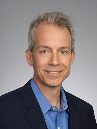 A man with short gray hair and blue eyes is wearing a navy suit jacket and a blue shirt, smiling at the camera against a plain gray background.