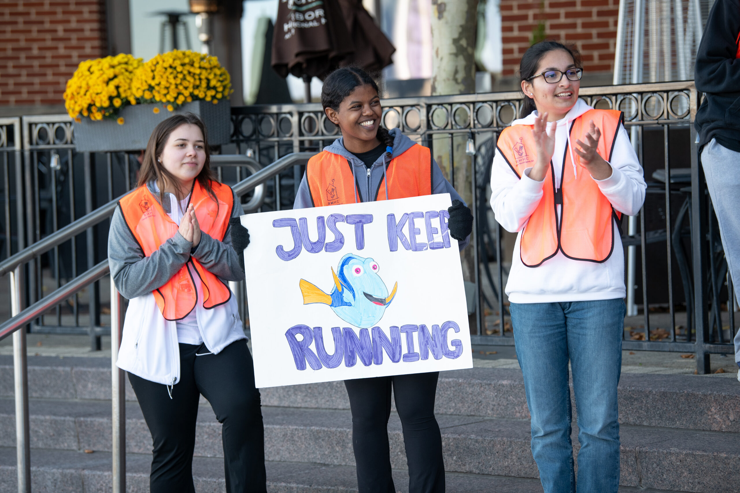 Three individuals wearing orange safety vests stand together. One holds a sign featuring a cartoon fish with the text "Just Keep Running," while the others clap. Yellow flowers are visible in the background.