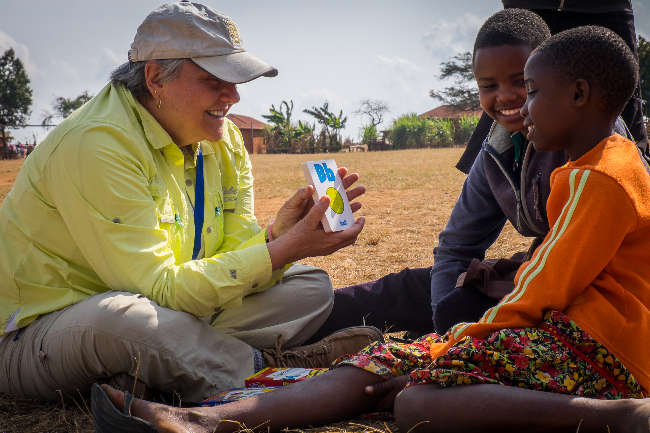 A person in a yellow shirt and hat shows flashcards to two children sitting on the ground, smiling and engaging with the activity. The setting appears to be an outdoor, rural area.
