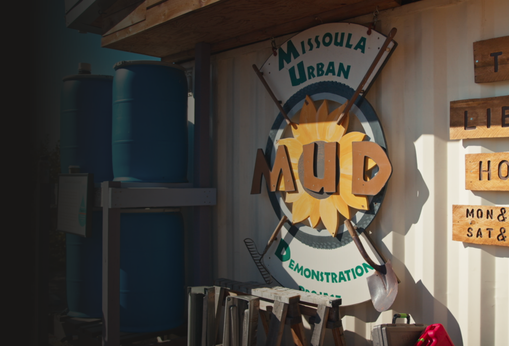 Outside view of the Missoula Urban Demonstration Project with large water tanks, gardening tools, and a wooden signboard featuring the acronym MUD.
