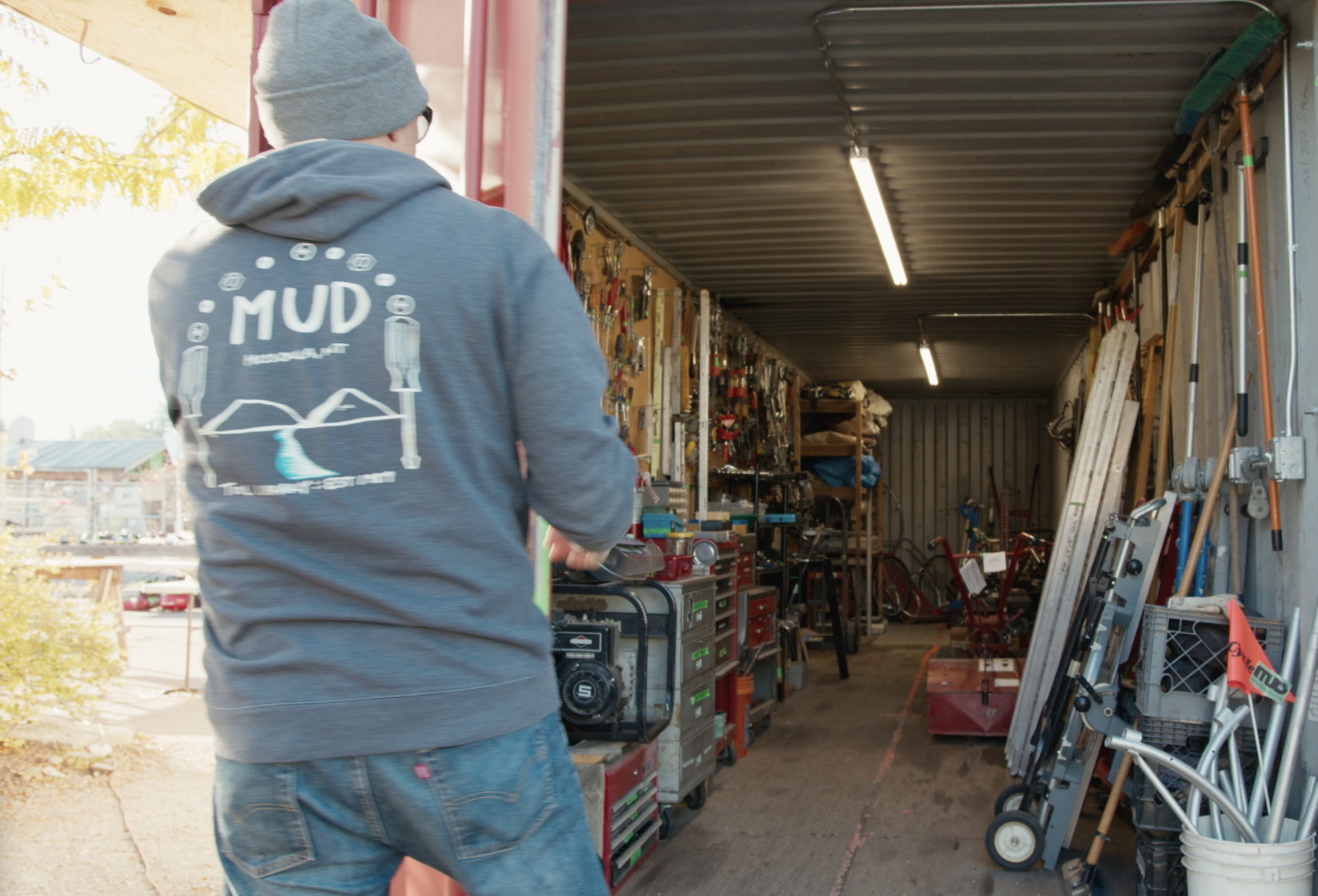 Person wearing a gray beanie and hoodie enters a storage container filled with various tools and equipment. The back of the hoodie says "MUD" along with a logo and additional text.