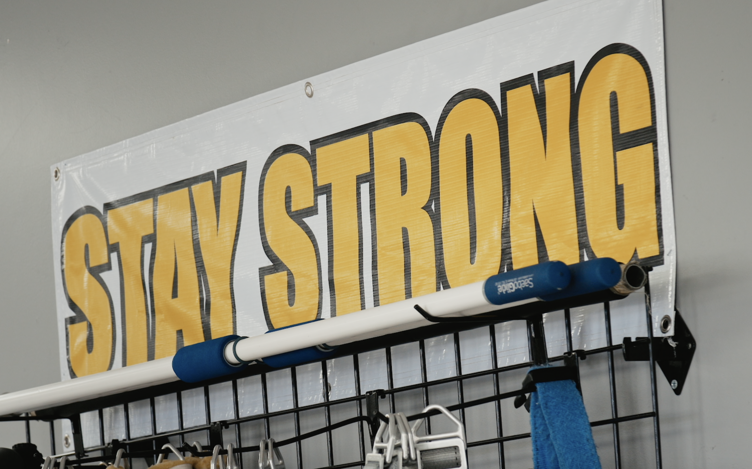 A "STAY STRONG" banner in yellow letters is pinned on a wall above a metal rack holding exercise equipment in a gym setting.