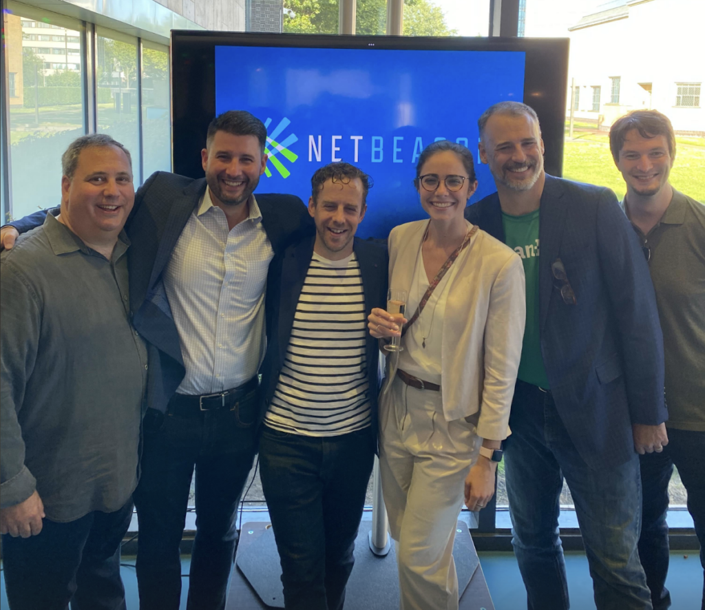 Six people standing together smiling in front of a screen displaying the logo "NetBeans." Four men and one woman wear business casual clothing. One man holds a glass and has his arm around another person.