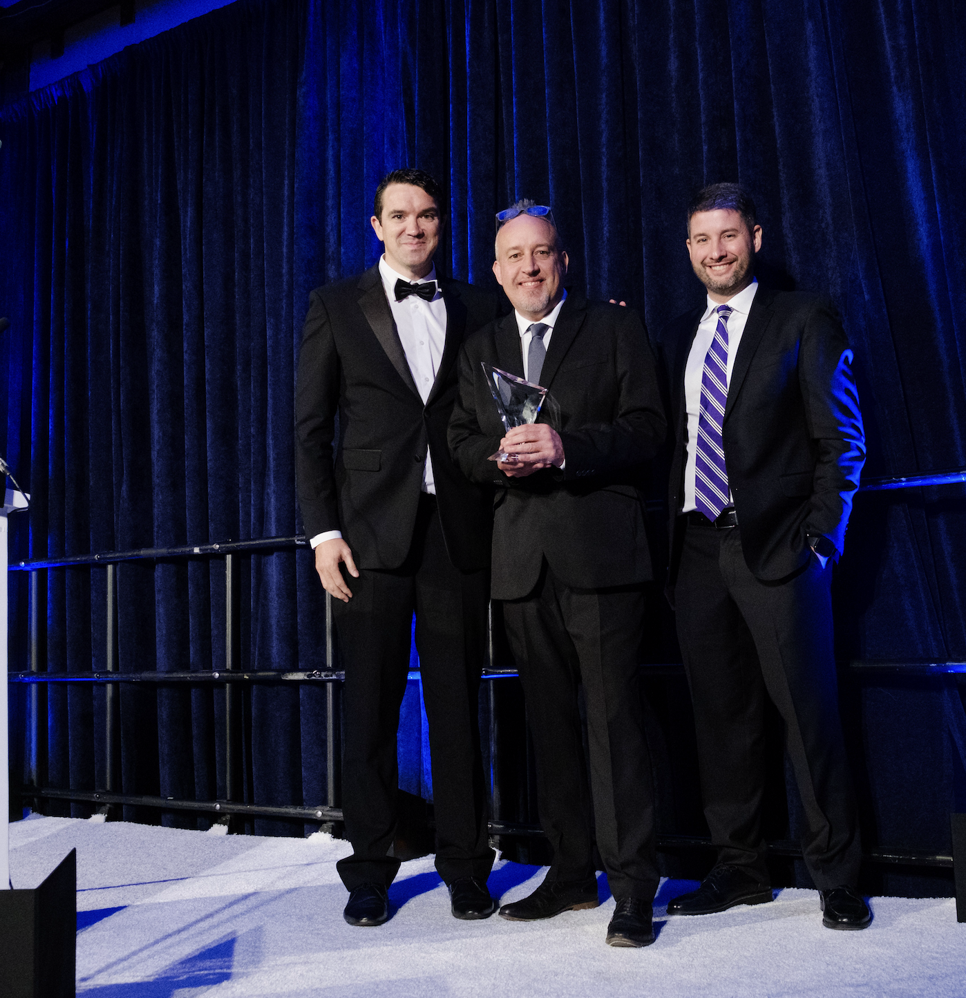 Three men in suits stand on a stage with dark blue curtains. The man in the center is holding a trophy, and the other two are standing on either side of him, smiling.